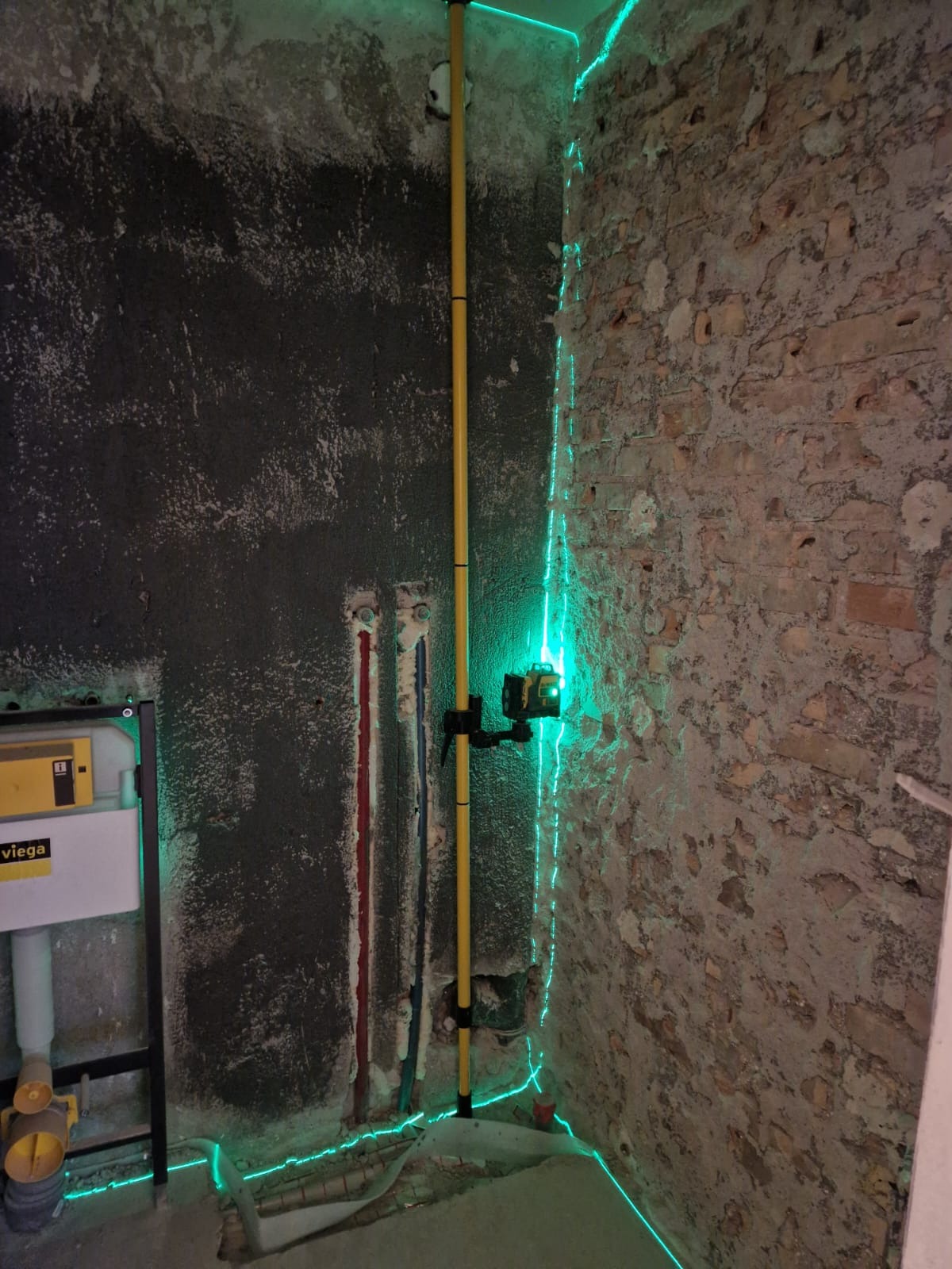 Surowa ściana z instalacją, poziomica laserowa wyznacza pion, widoczny stelaż podtynkowy WC Viega. Etap prac w remontowanej łazience, surowe wykończenie, ściana z cegły.