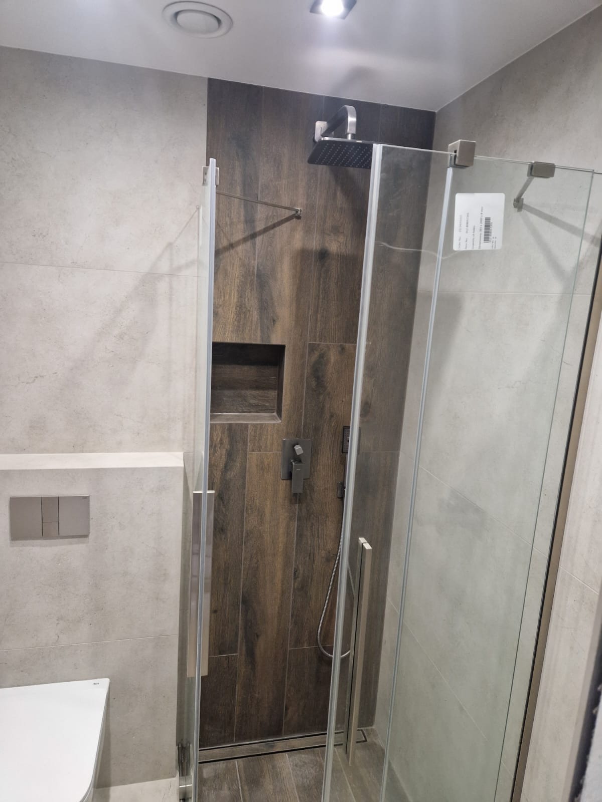 Nowoczesna kabina prysznicowa z deszczownicą i wnęką w ścianie wyłożonej płytkami imitującymi drewno, w łazience z szarymi płytkami.