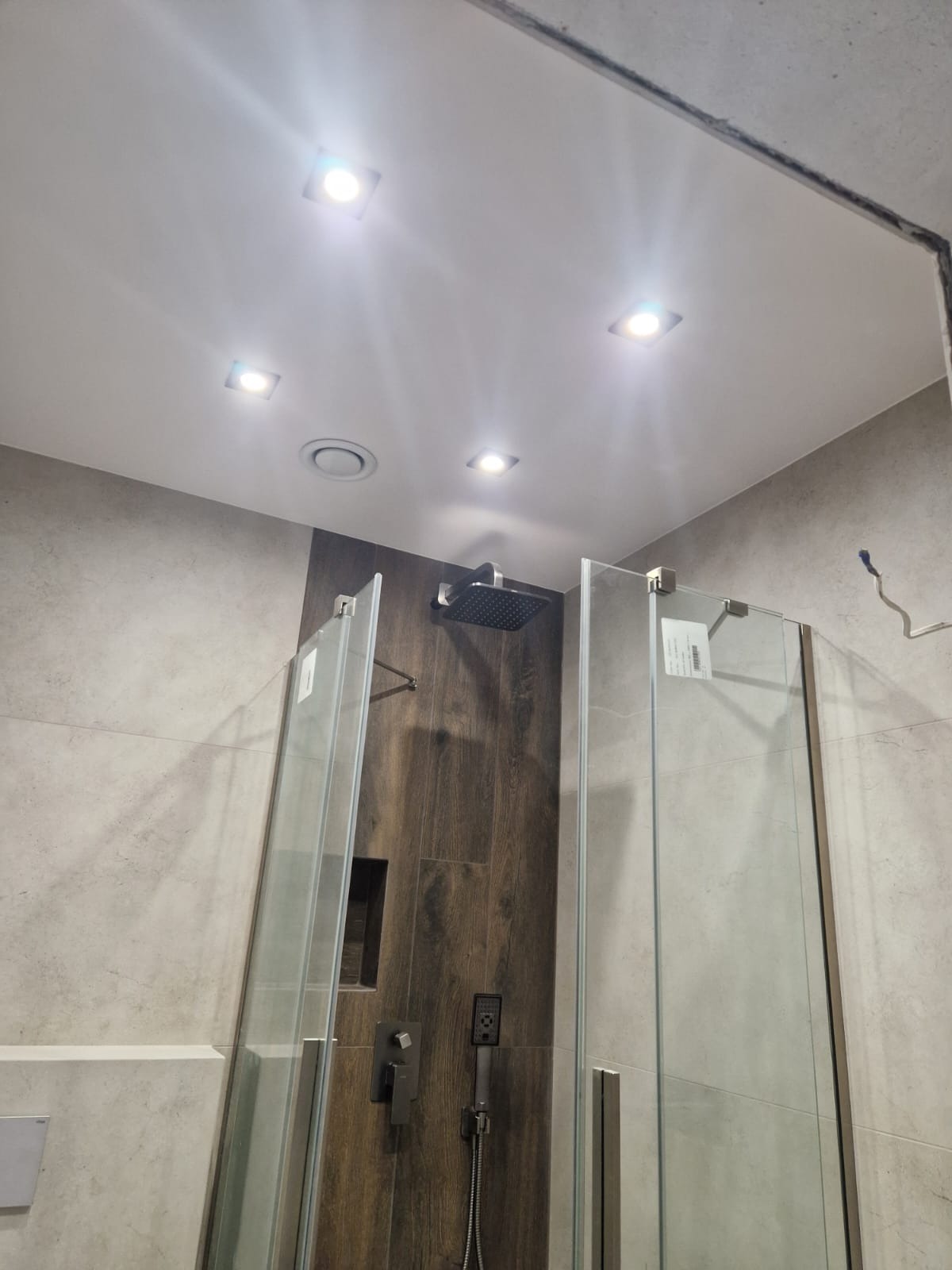 Nowoczesna kabina prysznicowa z deszczownicą i szklanymi drzwiami, wykończona płytkami w odcieniach szarości i brązu, z oświetleniem punktowym.