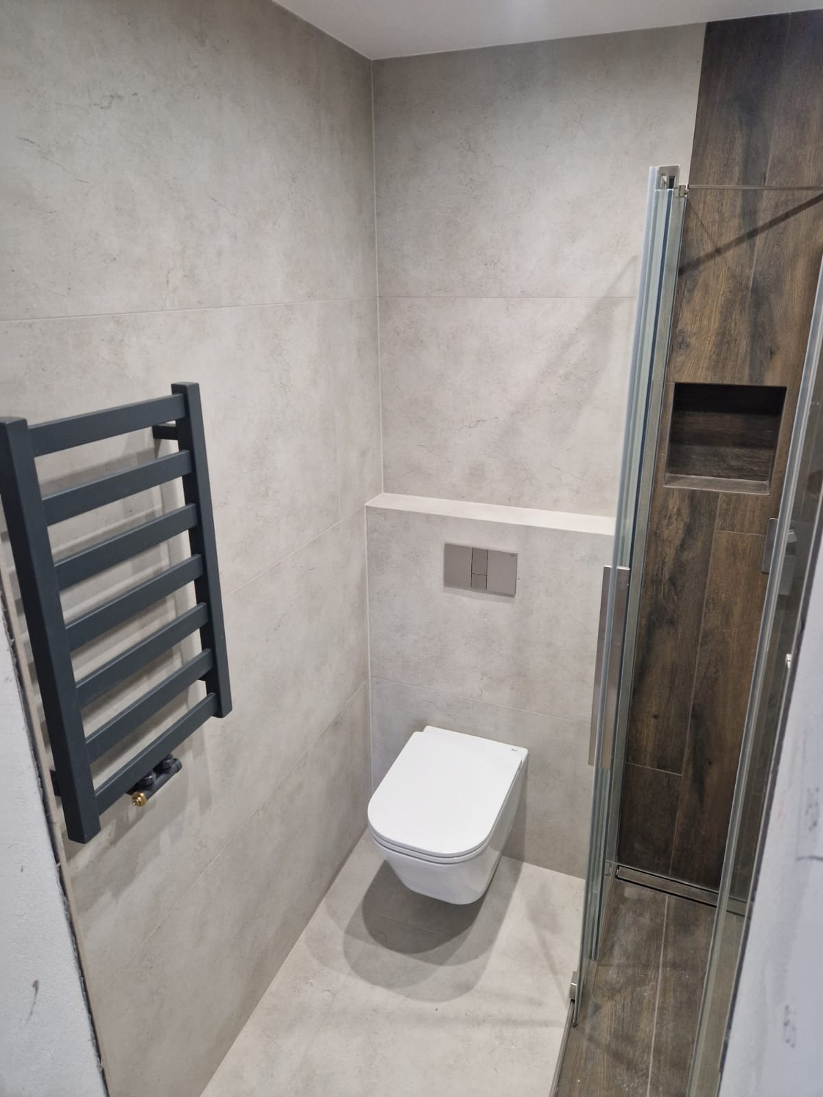 Nowoczesna łazienka z szarymi płytkami, grzejnikiem drabinkowym i kabiną prysznicową z drewnopodobnymi elementami. Uwagę zwraca minimalistyczna toaleta podtynkowa.