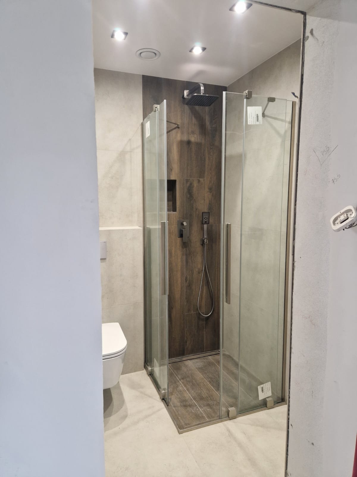 Nowoczesna kabina prysznicowa z deszczownicą i panelami imitującymi drewno, widok z częściowo wykończonej łazienki, z toaletą w kadrze.