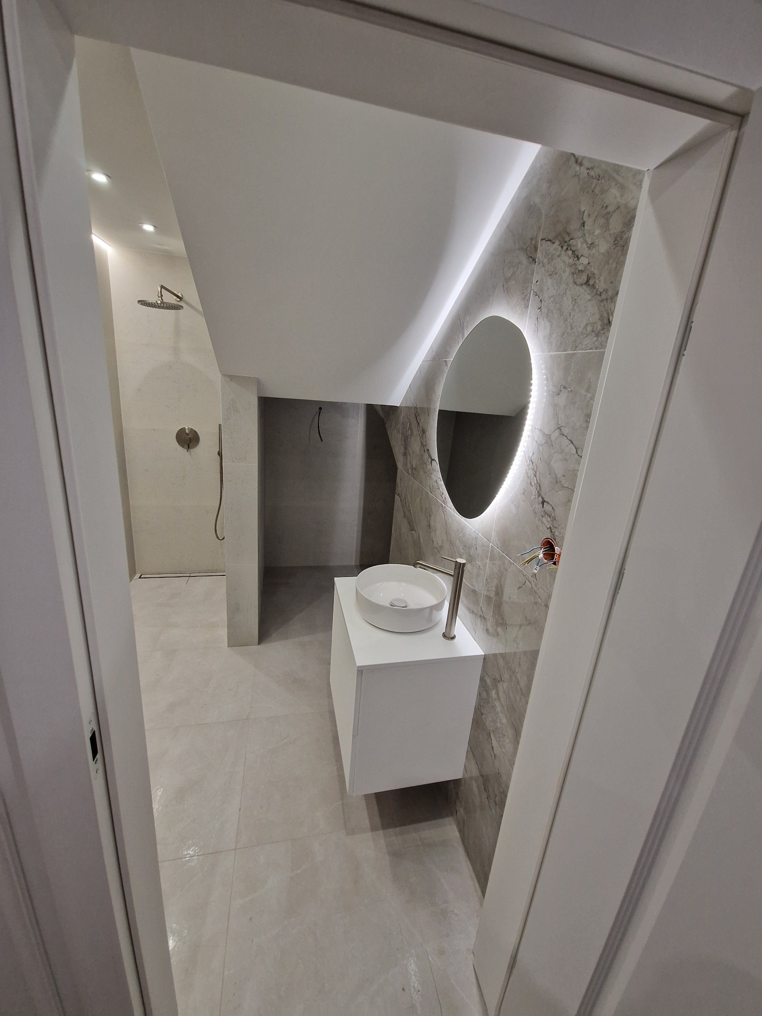 Nowoczesna łazienka w odcieniach szarości z owalnym lustrem podświetlanym LED, białą szafką z umywalką i kabiną prysznicową w tle, widziana zza uchylonych drzwi.