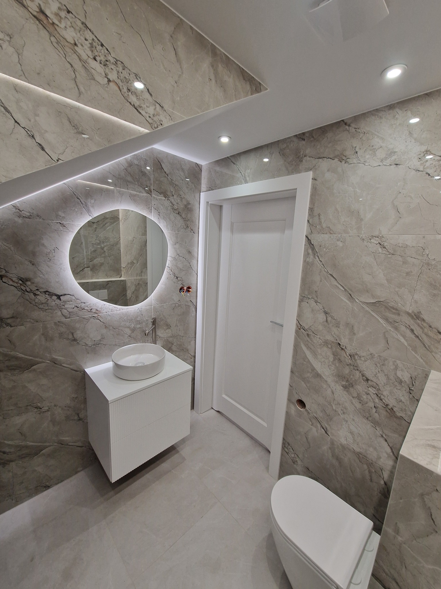 Wykończona łazienka z marmurowymi płytkami, podświetlanym lustrem i białą armaturą. Uwagę przykuwa owalne lustro z ledowym podświetleniem i minimalistyczna szafka pod umywalką.