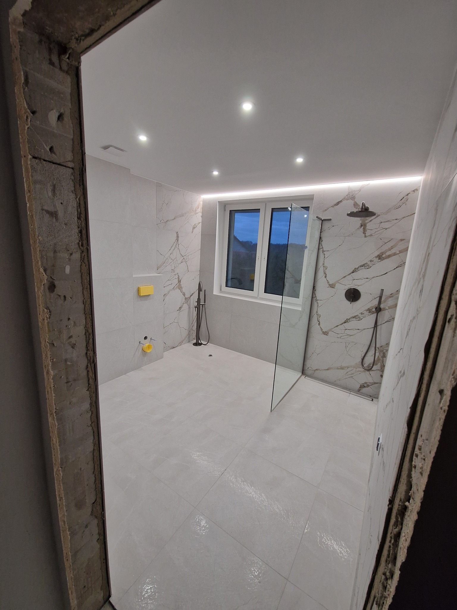 Wykończona łazienka z marmurowymi płytkami, kabiną prysznicową typu walk-in i oświetleniem LED, widziana zza surowej futryny drzwi.