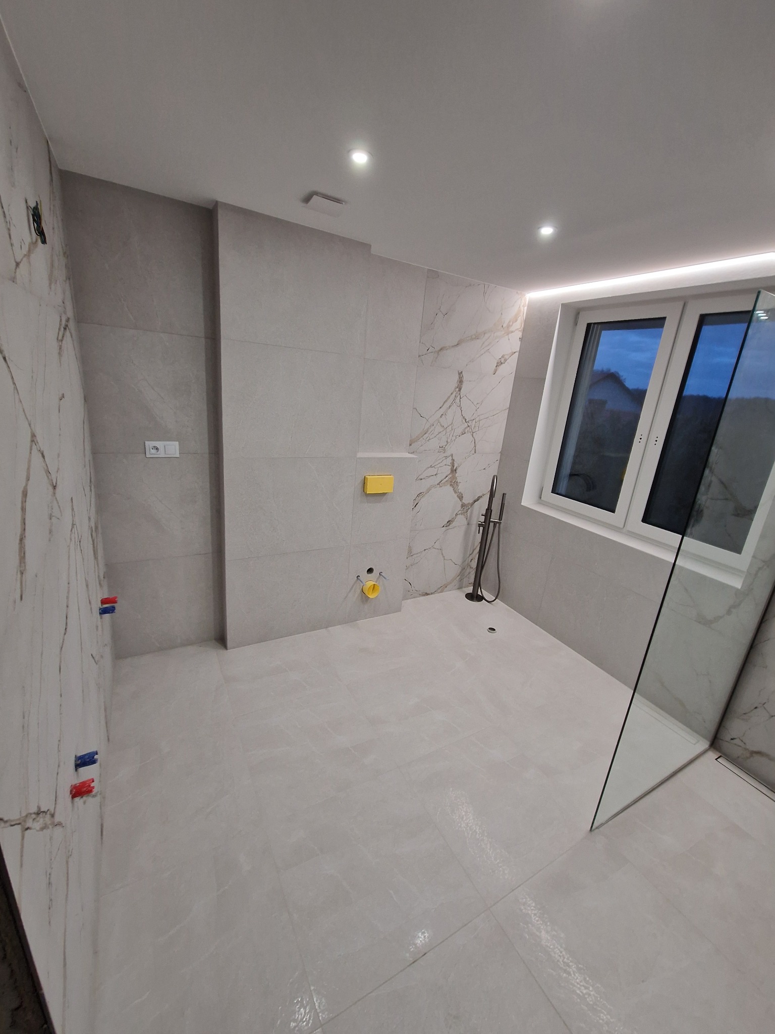 Wykończona łazienka z marmurowymi płytkami, szklaną kabiną prysznicową i oknem. Widoczne elementy instalacji i armatury w kolorze złotym. Nowoczesny design.