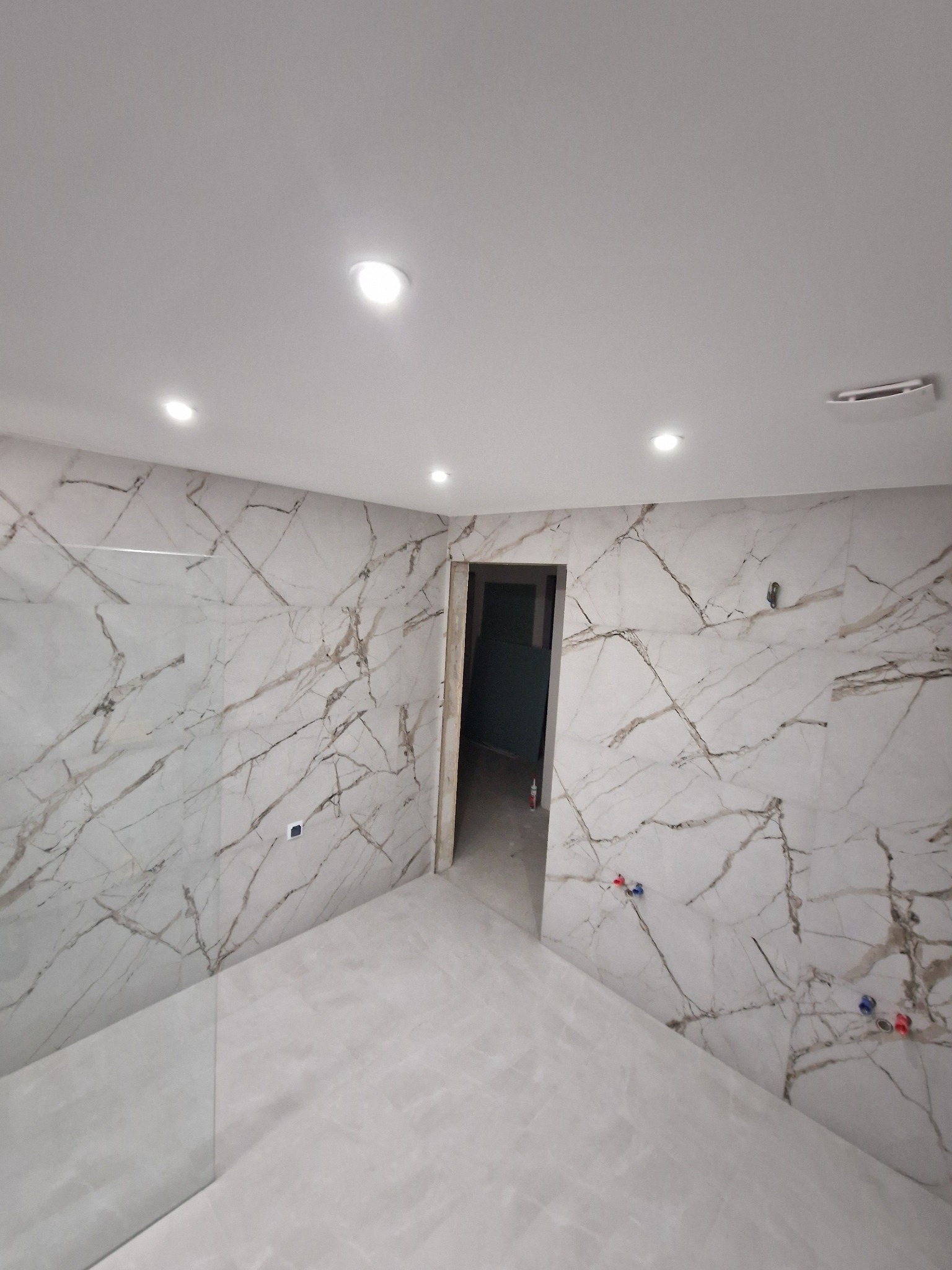 Wykończona łazienka z marmurowymi płytkami na ścianach i podłodze, oświetleniem punktowym i otwartym wejściem do innego pomieszczenia. Widoczne przyłącza wodne.