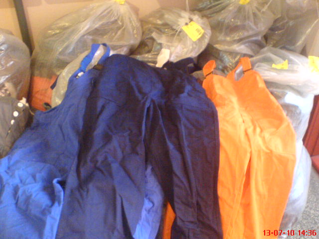 Stos niebieskich i pomarańczowych spodni ogrodniczek roboczych, zapakowanych w przezroczyste worki, widoczny fragment daty na zdjęciu.