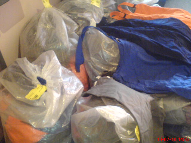 Używana odzież robocza, w tym spodnie ogrodniczki, zapakowana w przezroczyste worki foliowe z żółtymi etykietami, widoczne także inne elementy odzieży w kolorach pomarańczowym, niebieskim i szarym.