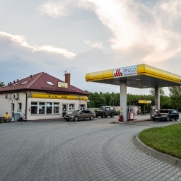 ARCAB Investments Sp. z o.o. - Stacja paliw z budynkiem motelowym i barem, dwa samochody tankują, dwóch mężczyzn siedzi na ławce przed budynkiem, pochmurne niebo.