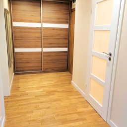 ARCAB Investments Sp. z o.o. - Jasne panele podłogowe w korytarzu z przesuwną szafą z drewnianymi frontami i białymi pasami oraz białe drzwi z przeszkleniami.