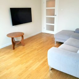 ARCAB Investments Sp. z o.o. - Jasne wnętrze salonu z drewnianą podłogą, szarą sofą, telewizorem na ścianie i okrągłym stolikiem kawowym z drewna.