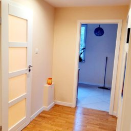 ARCAB Investments Sp. z o.o. - Jasny korytarz z drewnianą podłogą, białe drzwi z przeszkleniami i widok na pomieszczenie z mopem i dekoracyjną kulą, oświetlenie punktowe