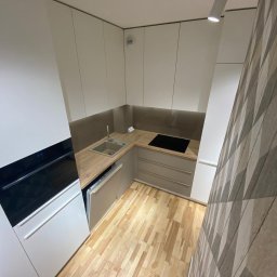 ARCAB Investments Sp. z o.o. - Nowoczesna, minimalistyczna kuchnia z białymi szafkami, drewnianym blatem i podłogą, oświetlona punktowymi lampami sufitowymi, z dekoracyjną tapetą w geometryczne wzory na ścianie.