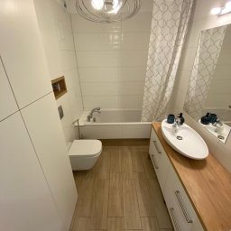 ARCAB Investments Sp. z o.o. - Jasna łazienka z drewnianym blatem umywalki, białą armaturą i podłogą z płytek imitujących drewno. W tle wanna z zasłoną w geometryczne wzory.