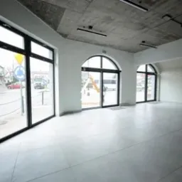 Puste, jasne pomieszczenie z dużymi oknami wychodzącymi na ulicę z ruchem, szary betonowy sufit z zamontowanymi lampami, białe ściany i szara podłoga z polerowanego betonu.