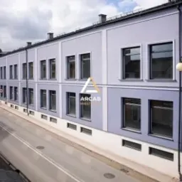 Dwupiętrowy budynek biurowy z szarymi ścianami i rzędem okien, z logo firmy na elewacji, widok z lekkiej góry na ulicę z zaparkowanymi samochodami.