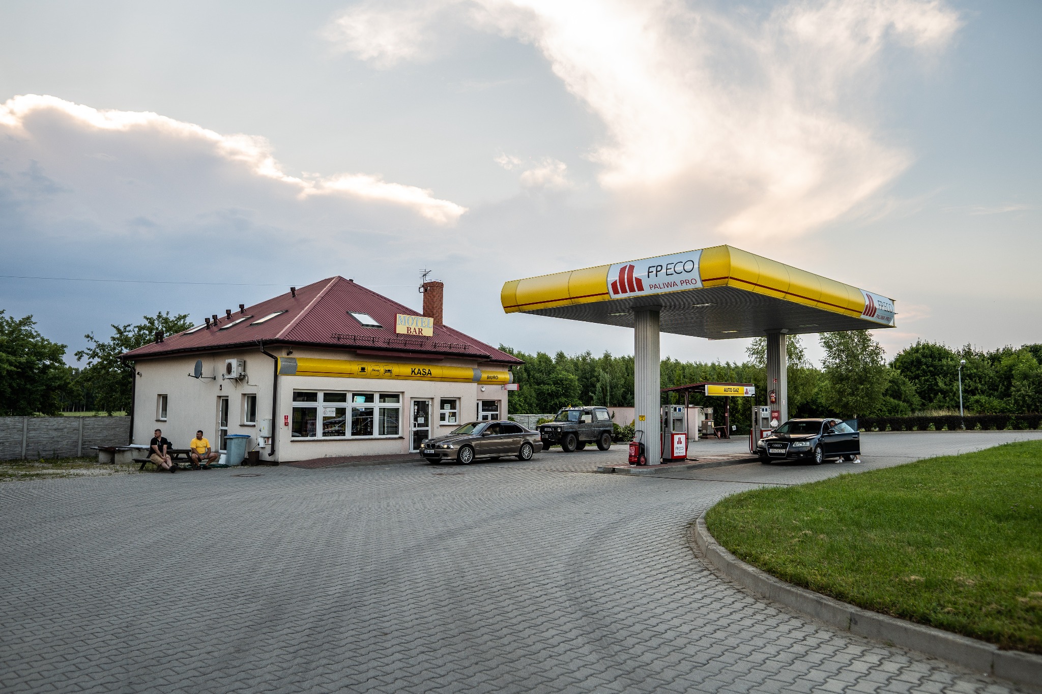 Stacja paliw z budynkiem motelowym i barem, dwa samochody tankują, dwóch mężczyzn siedzi na ławce przed budynkiem, pochmurne niebo.