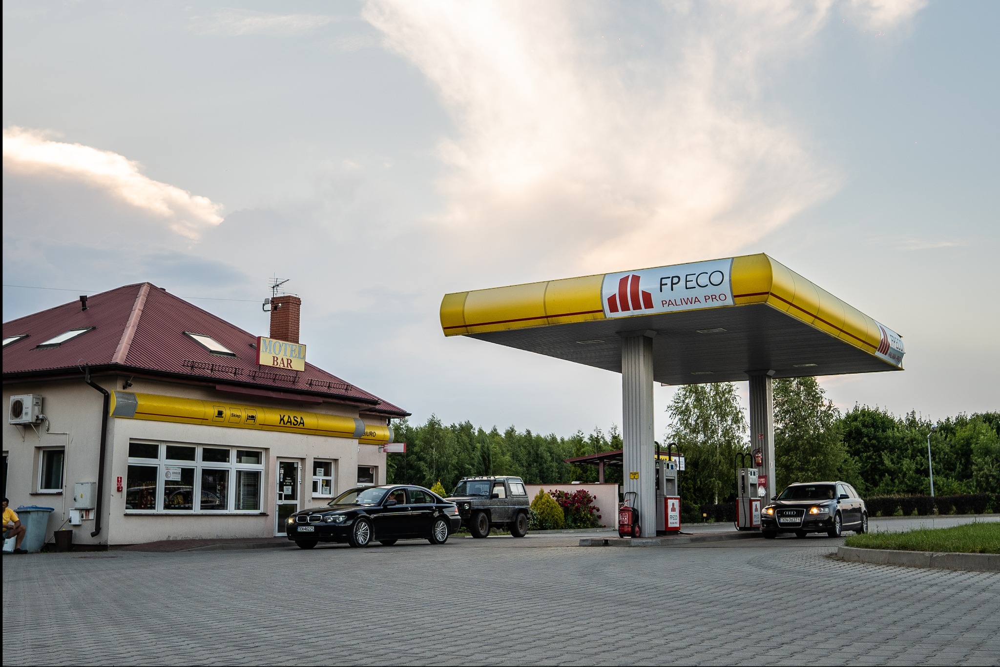 Stacja paliw FP ECO z budynkiem motel-baru w tle, kilka samochodów na brukowanym placu przed dystrybutorami i budynkiem, wieczorne oświetlenie.