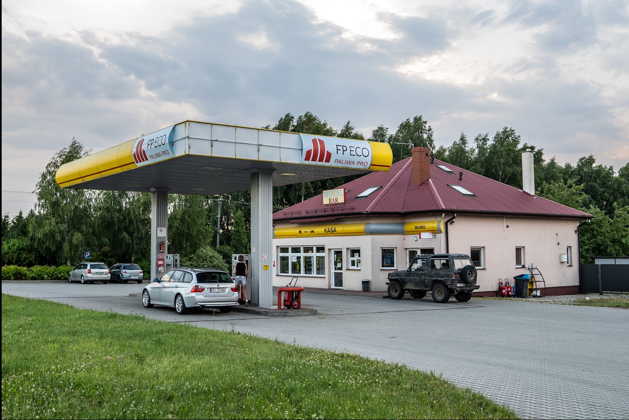 Stacja paliw z podjazdem, dystrybutorami, budynkiem z napisem 'KASA', 'BIURO' i 'MOTEL BAR', zaparkowanymi samochodami osobowymi i terenowym.