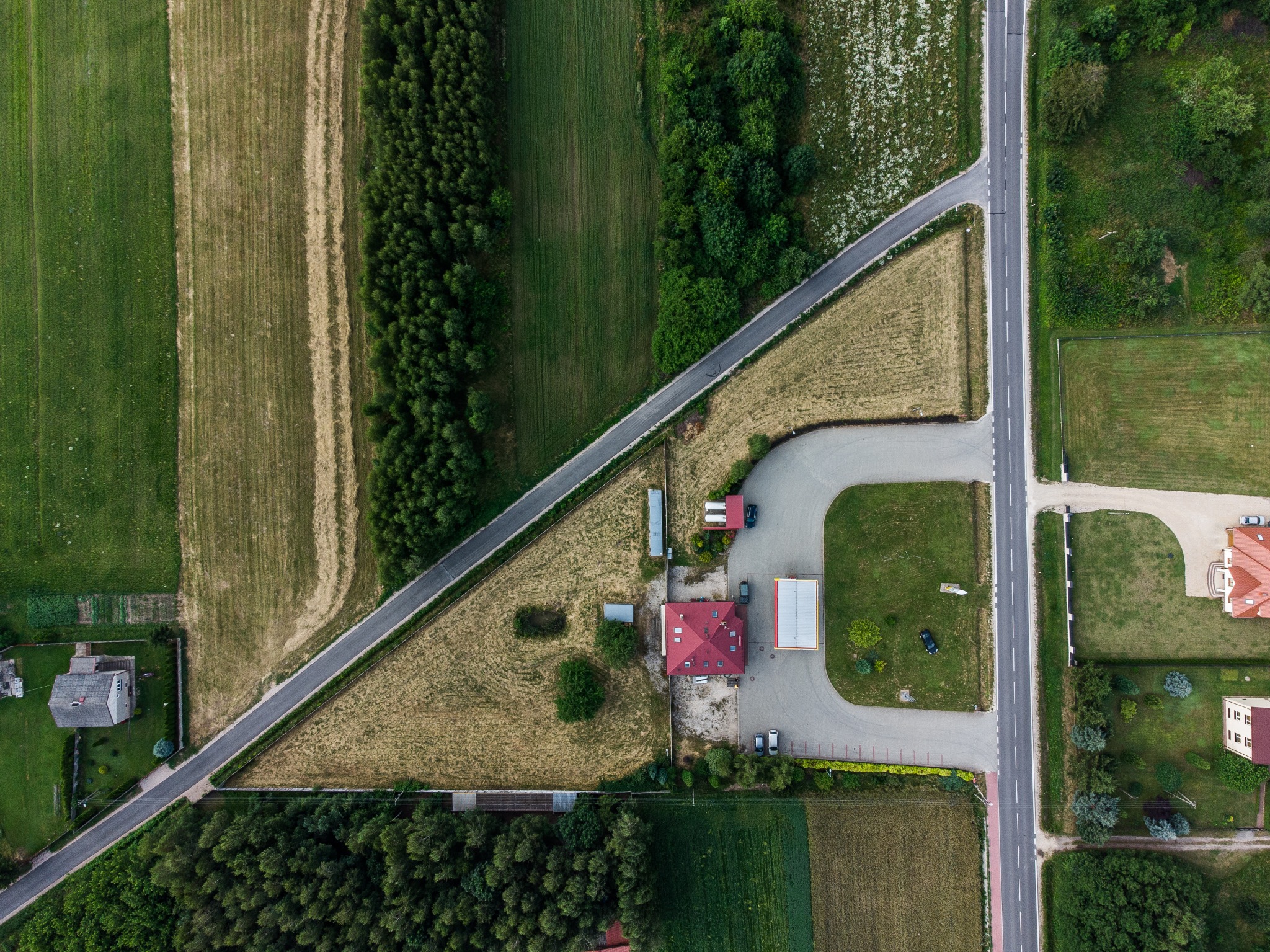 Widok z lotu ptaka na skrzyżowanie dróg wiejskich, pola uprawne i zabudowania, w tym dom z czerwoną dachówką oraz budynek gospodarczy z szarą blachą falistą, otoczone zielenią i parkingiem...