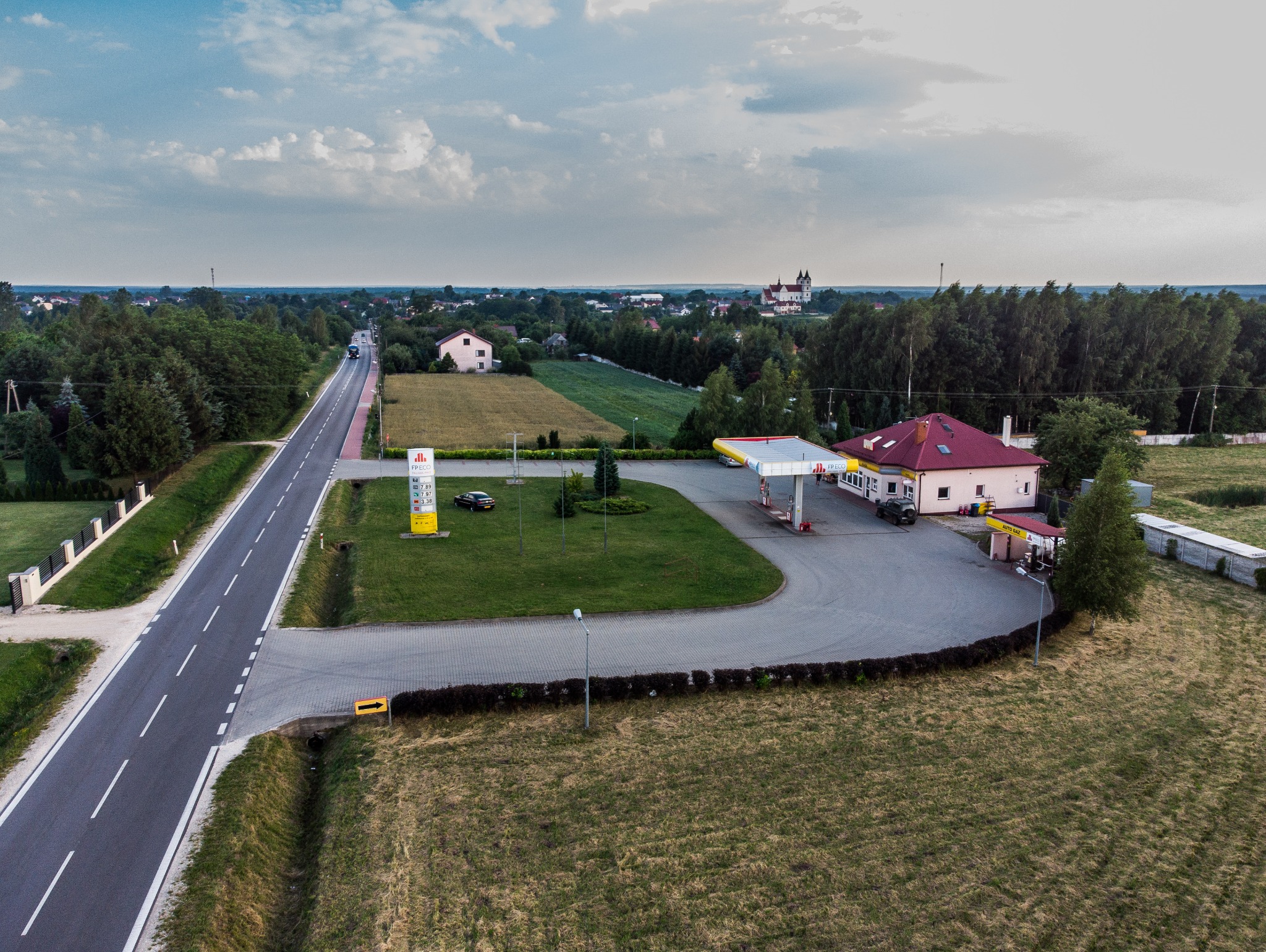 Panorama stacji benzynowej z budynkiem w kolorze beżowym i czerwonym dachem, widok z lotu ptaka, w tle wieś z kościołem.