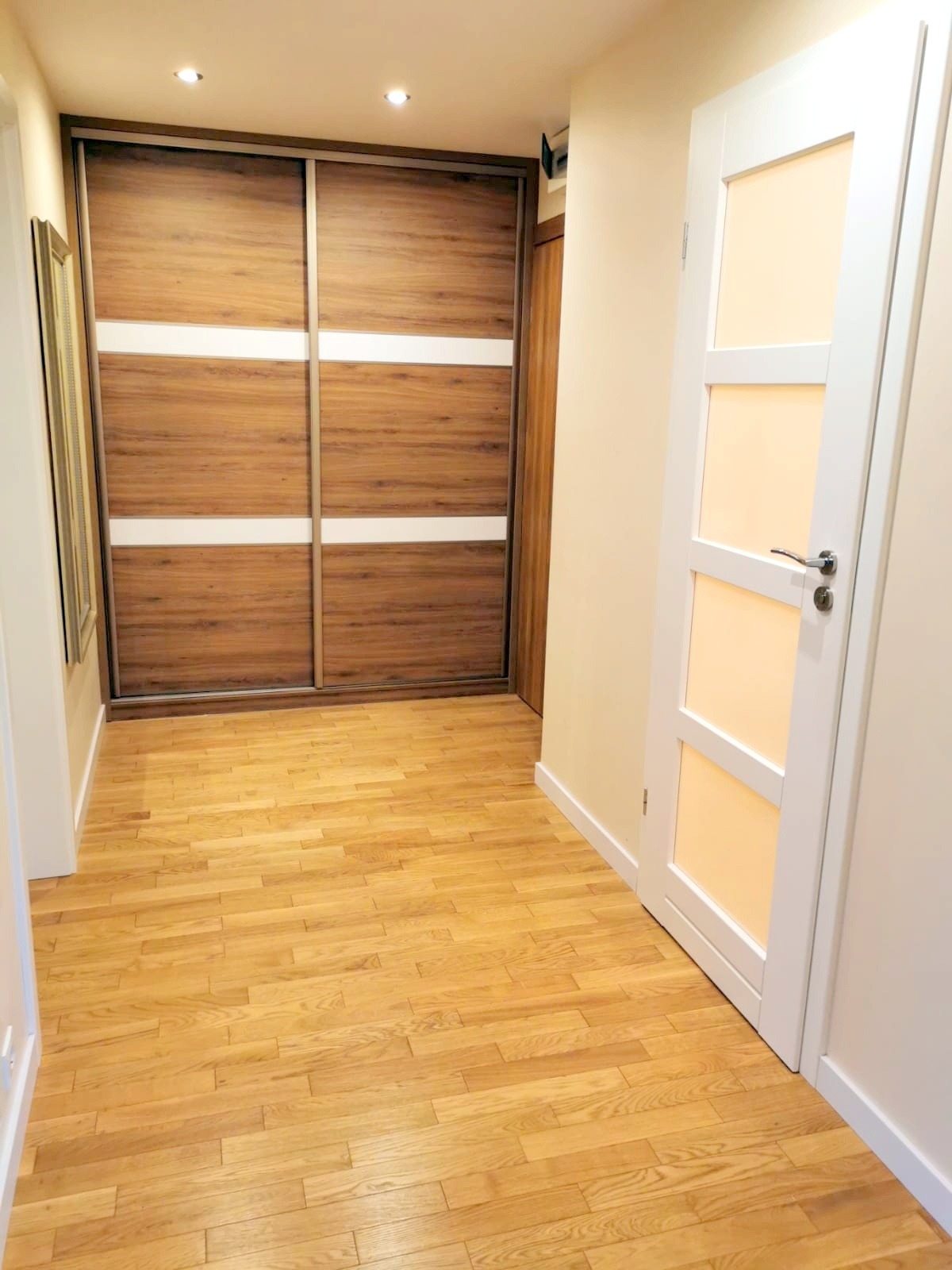 Jasne panele podłogowe w korytarzu z przesuwną szafą z drewnianymi frontami i białymi pasami oraz białe drzwi z przeszkleniami.