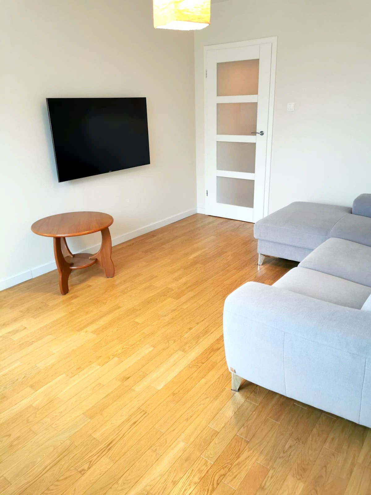 Jasne wnętrze salonu z drewnianą podłogą, szarą sofą, telewizorem na ścianie i okrągłym stolikiem kawowym z drewna.