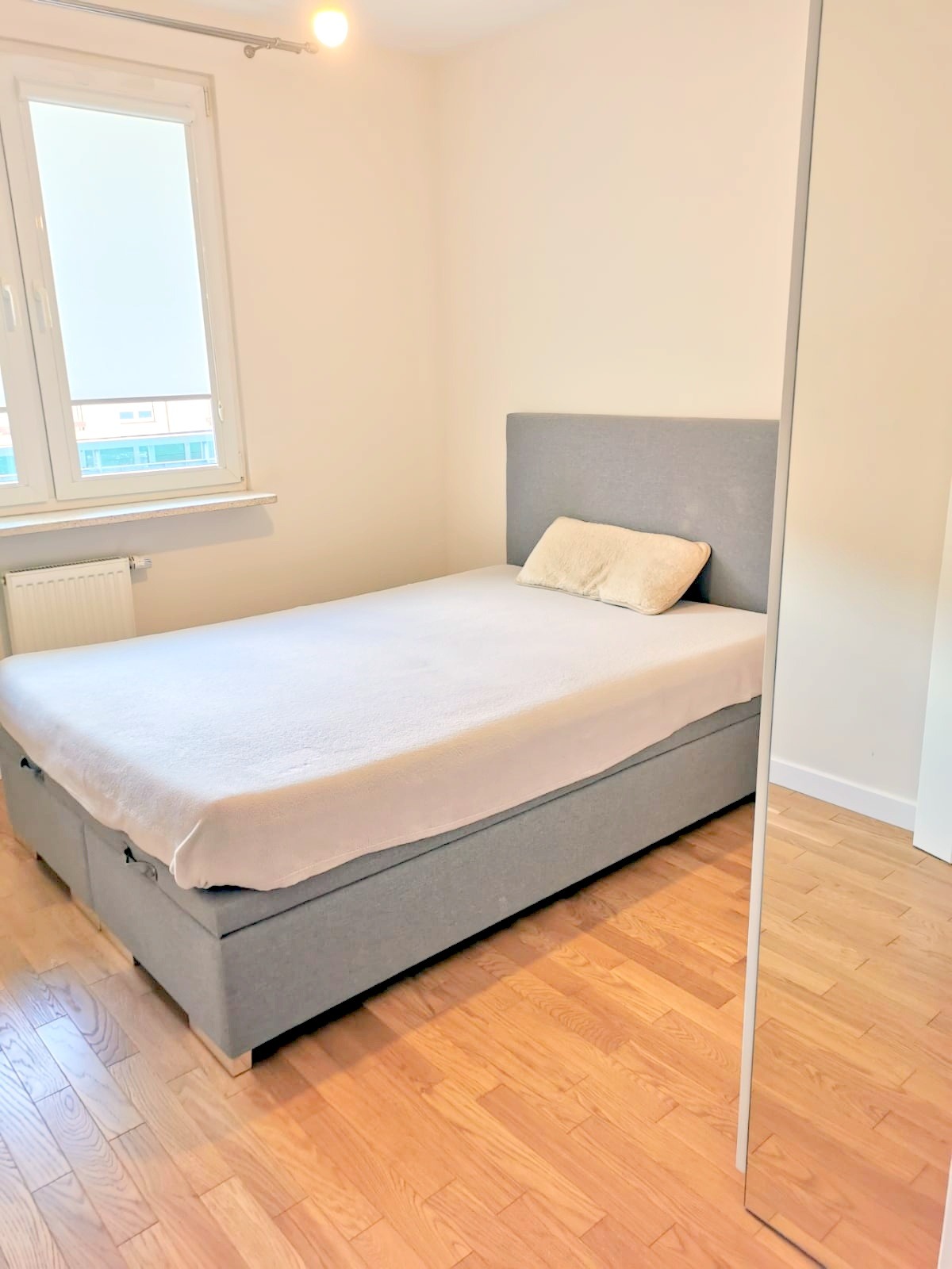 Jasne wnętrze sypialni z łóżkiem kontynentalnym w szarym obiciu, drewniana podłoga, okno z roletą w tle.