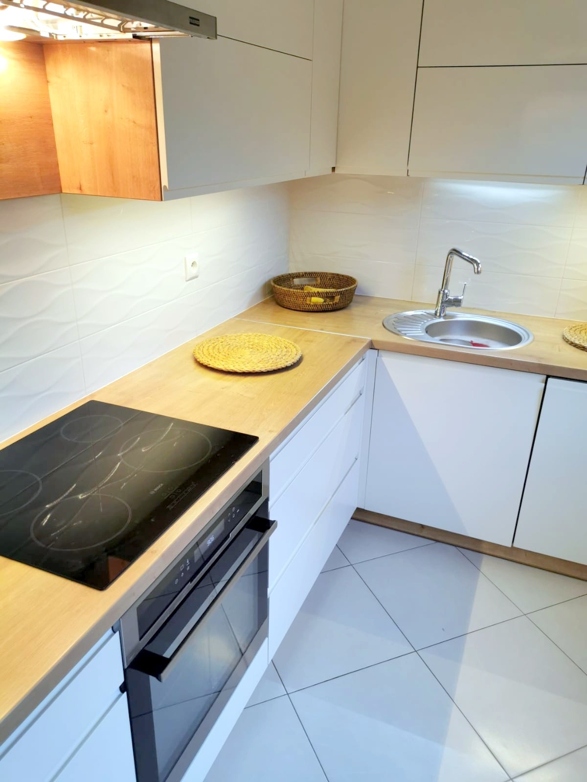 Nowoczesna kuchnia z białymi szafkami, drewnianym blatem, płytą indukcyjną i piekarnikiem, uzupełniona o pleciony koszyk i podkładki na blacie.