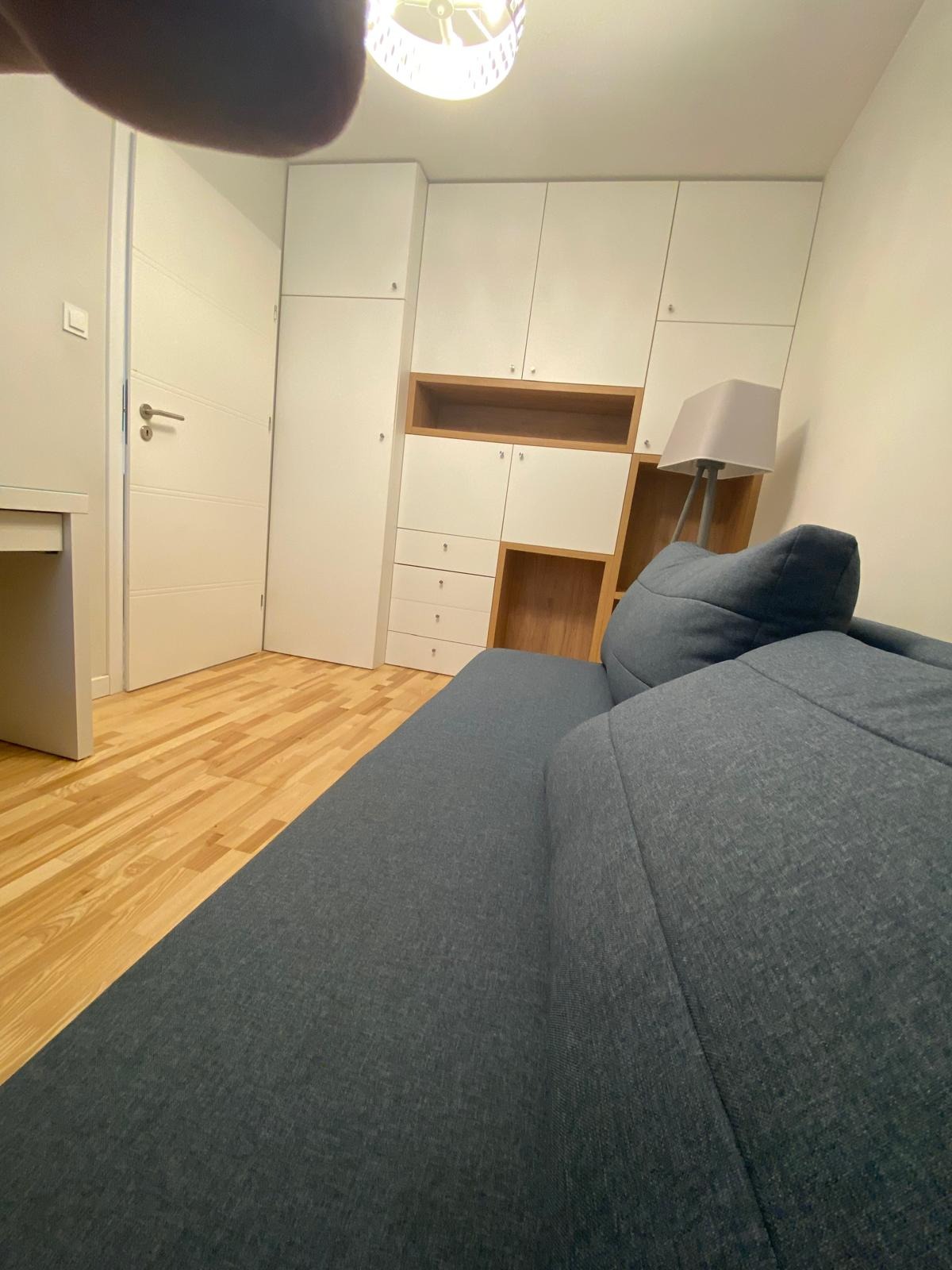 Wnętrze pokoju z niebieską sofą na pierwszym planie, widoczna zabudowa meblowa z białymi szafkami i drewnianymi wnękami, parkiet na podłodze i lampa podłogowa.