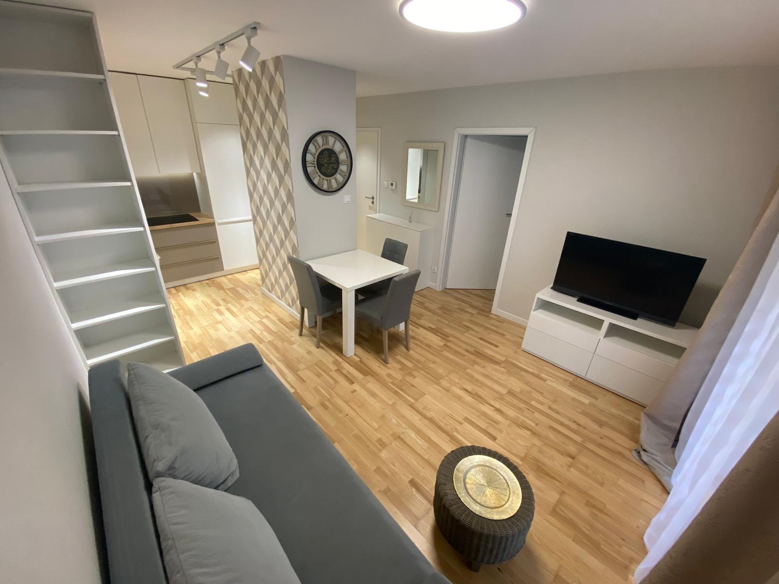 Jasne wnętrze mieszkania z drewnianą podłogą, białymi meblami kuchennymi z półkami, stołem z krzesłami, szarą sofą i telewizorem na komodzie.