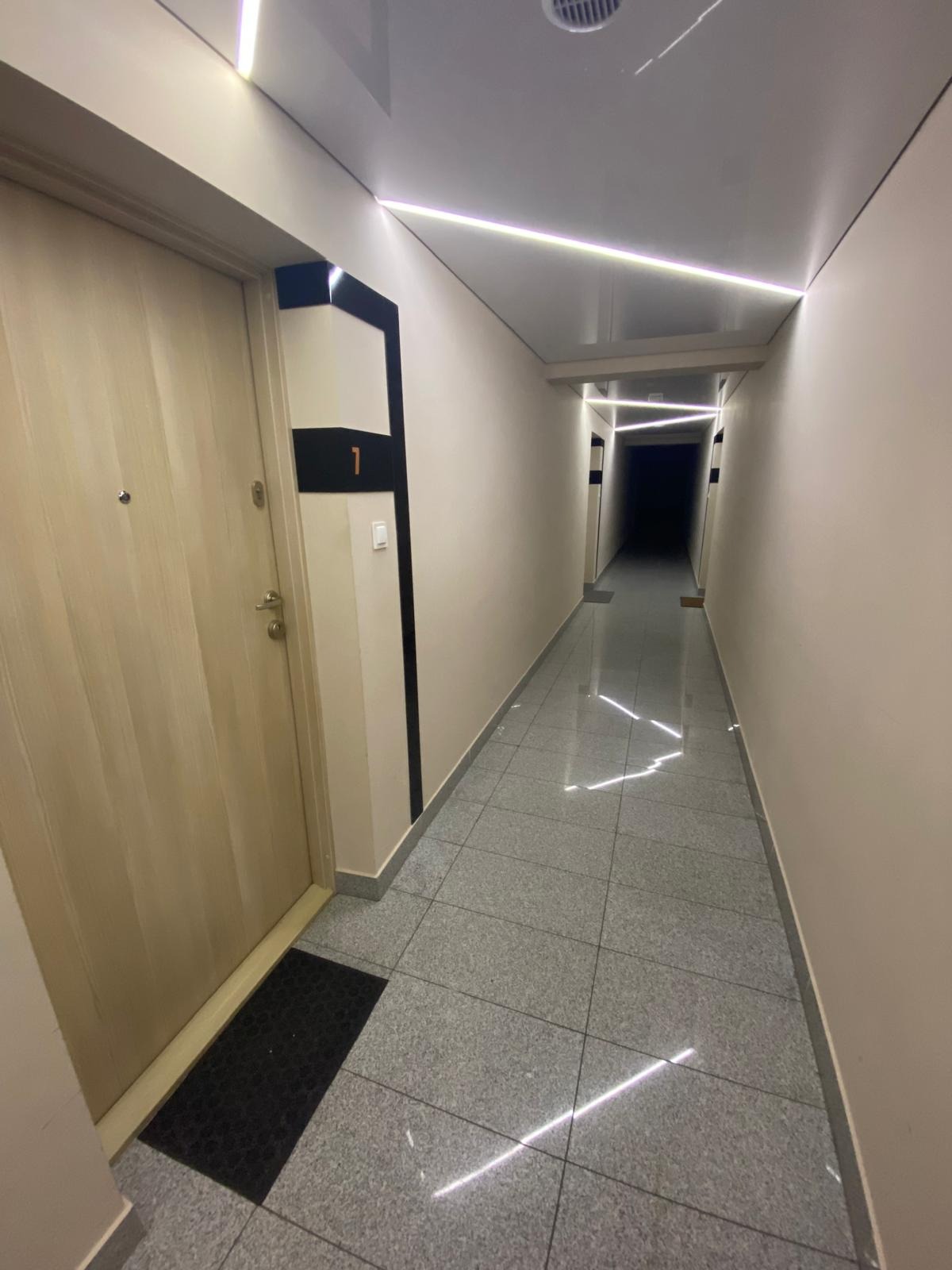Korytarz w apartamentowcu z numerem '1' na drzwiach i nowoczesnym oświetleniem LED na suficie, szara podłoga z płytek.