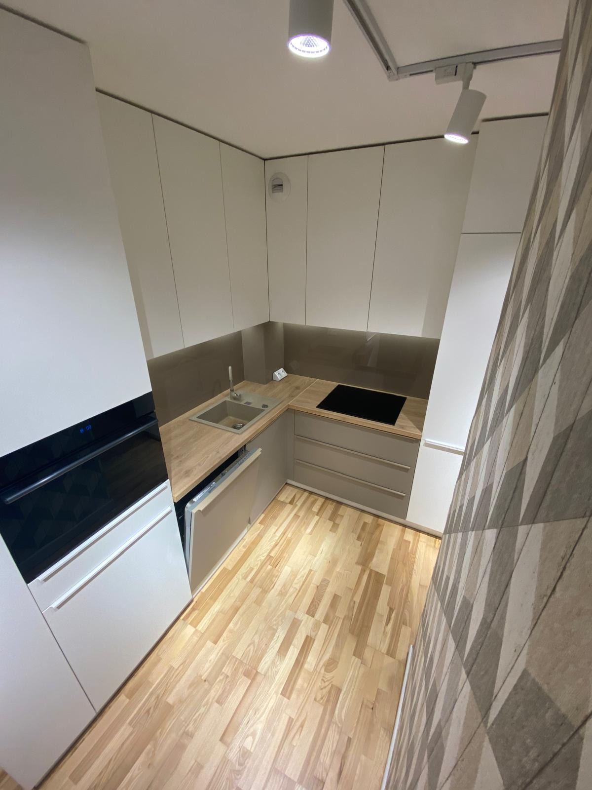 Nowoczesna, minimalistyczna kuchnia z białymi szafkami, drewnianym blatem i podłogą, oświetlona punktowymi lampami sufitowymi, z dekoracyjną tapetą w geometryczne wzory na ścianie.