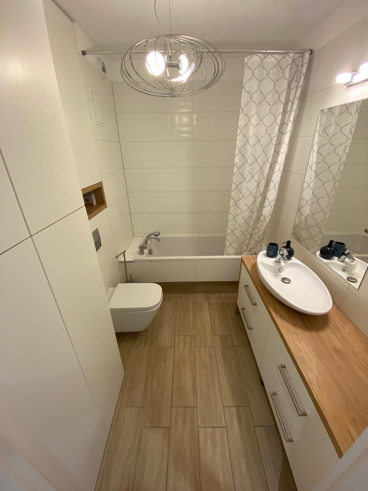 Jasna łazienka z drewnianym blatem umywalki, białą armaturą i podłogą z płytek imitujących drewno. W tle wanna z zasłoną w geometryczne wzory.