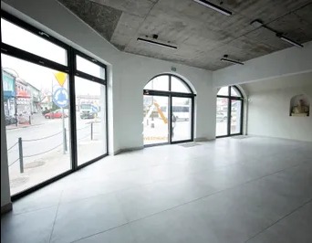 Puste, jasne pomieszczenie z dużymi oknami wychodzącymi na ulicę z ruchem, szary betonowy sufit z zamontowanymi lampami, białe ściany i szara podłoga z polerowanego betonu.