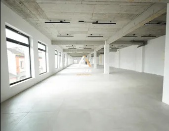 Przestronne, puste pomieszczenie biurowe w stanie deweloperskim z surowym betonowym sufitem, dużymi oknami po lewej stronie i betonową podłogą. W tle widoczny korytarz z kolumnami i logo firmy...