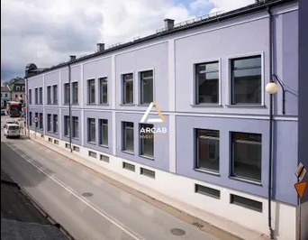 Dwupiętrowy budynek biurowy z szarymi ścianami i rzędem okien, z logo firmy na elewacji, widok z lekkiej góry na ulicę z zaparkowanymi samochodami.