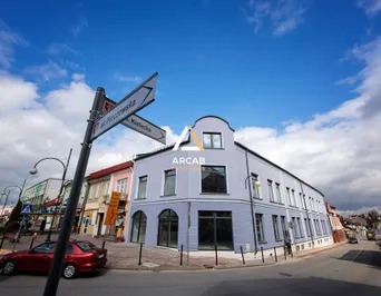 Narożny, dwupiętrowy budynek w jasnych barwach, z zaokrąglonym oknem na poddaszu, widoczny z ulicy z czerwonym samochodem i drogowskazem na pierwszym planie, błękitne niebo z chmurami w tle.