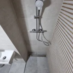 Nowoczesna kabina prysznicowa z deszczownicą, odpływem liniowym i płytkami w odcieniach szarości oraz dekoracyjną ścianą z drewnianymi lamelami. Widoczna umywalka po lewej stronie.