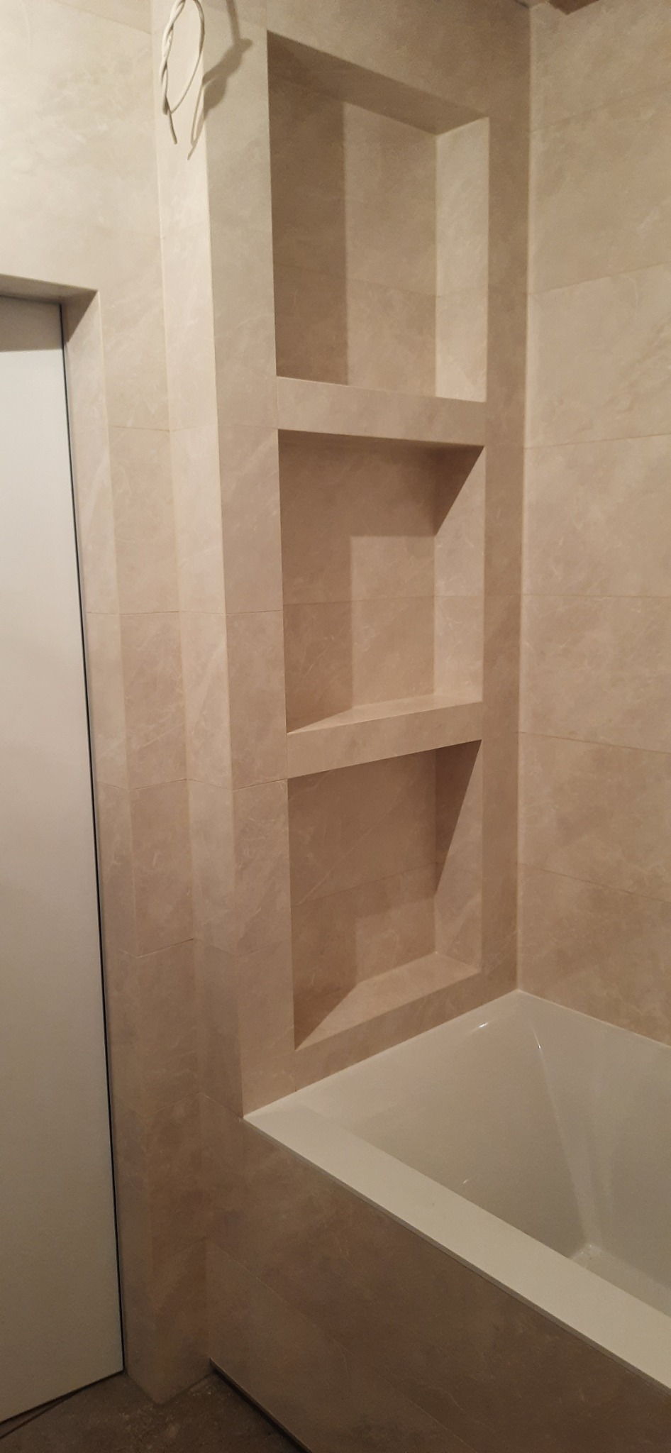 Beżowa łazienka z wanną i wnękami wyłożonymi płytkami imitującymi marmur, widoczne wystające przewody elektryczne