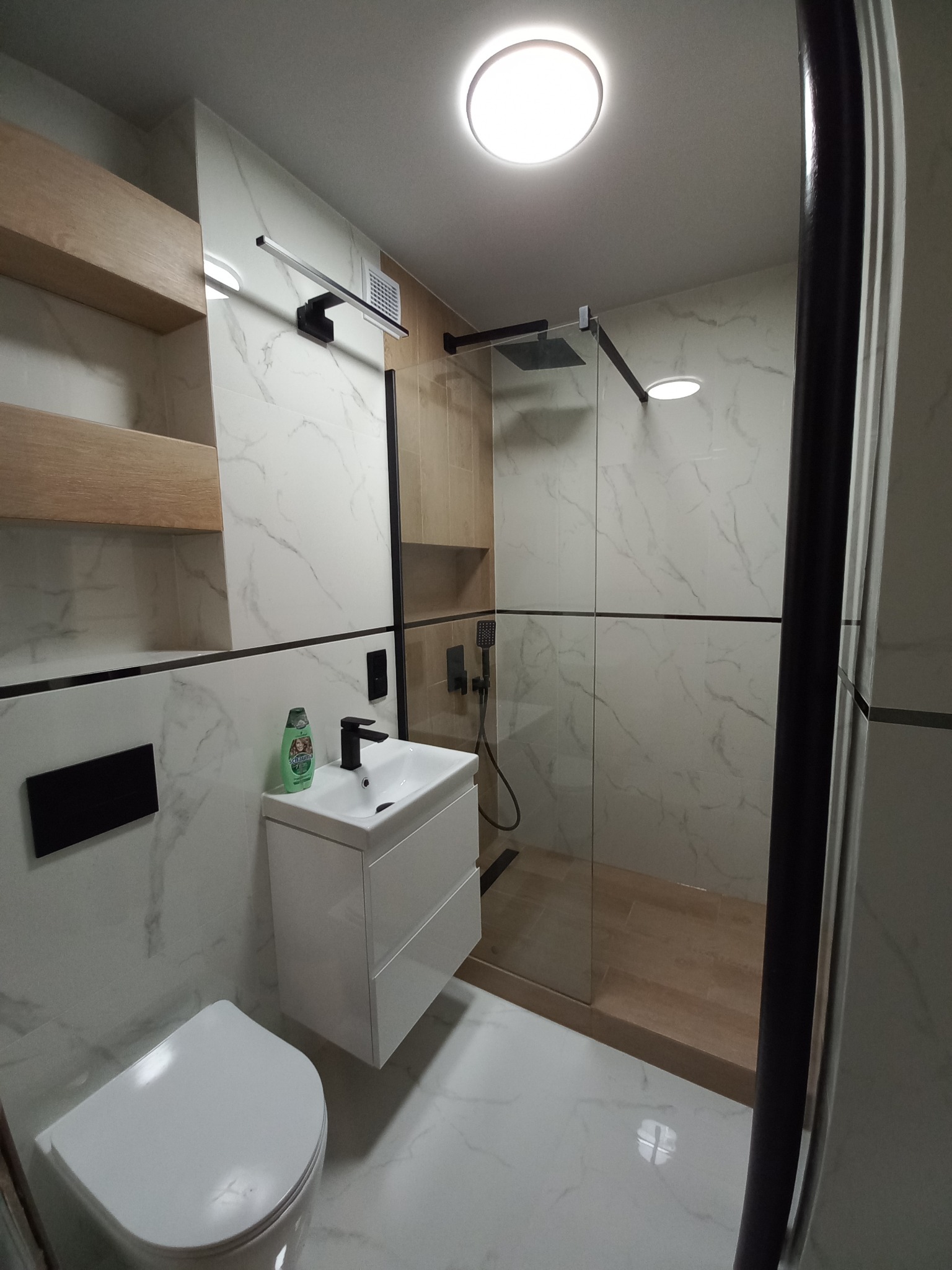 Nowoczesna łazienka z kabiną prysznicową typu walk-in, białymi płytkami imitującymi marmur, wiszącą szafką z umywalką, toaletą i drewnianymi półkami.