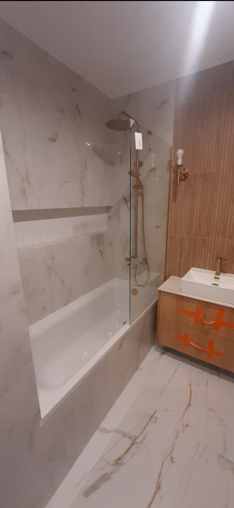 Nowoczesna łazienka z wanną za szklaną ścianką, deszczownicą w kolorze miedzi, umywalką nablatową i szafką z pomarańczowymi uchwytami na tle płytek imitujących marmur.