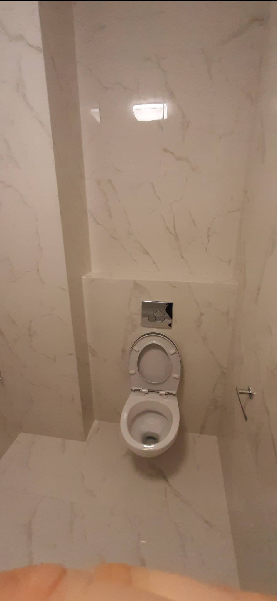 Toaleta wisząca z otwartą deską, chromowany przycisk spłuczki, płytki imitujące marmur na ścianach i podłodze, widok z góry.