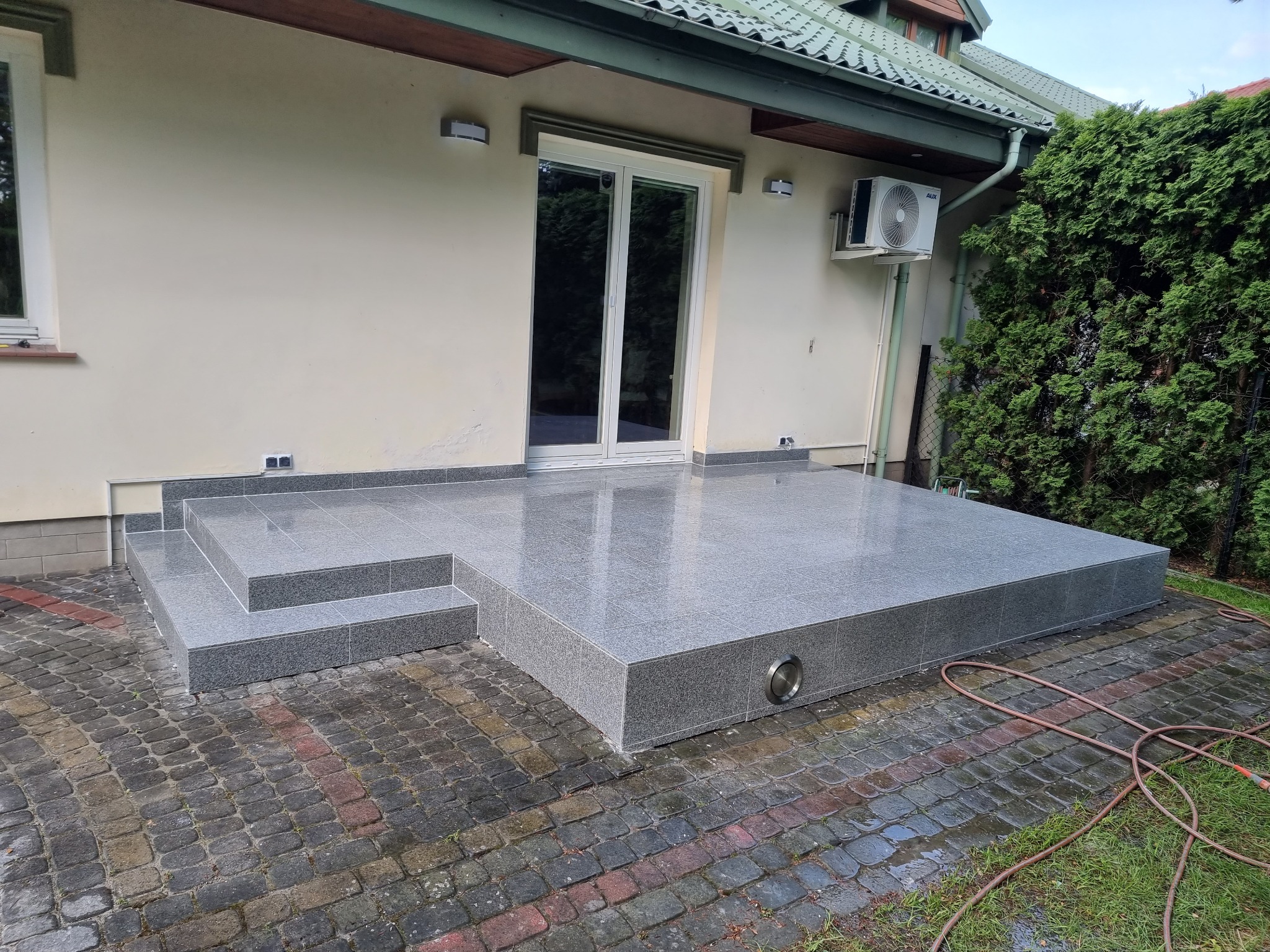 Wykończony taras z szarych płytek granitowych z trzema stopniami, widoczne oświetlenie wbudowane w obrzeże tarasu, obok posesji z zielonymi tujami, na zewnątrz budynku z klimatyzatorem