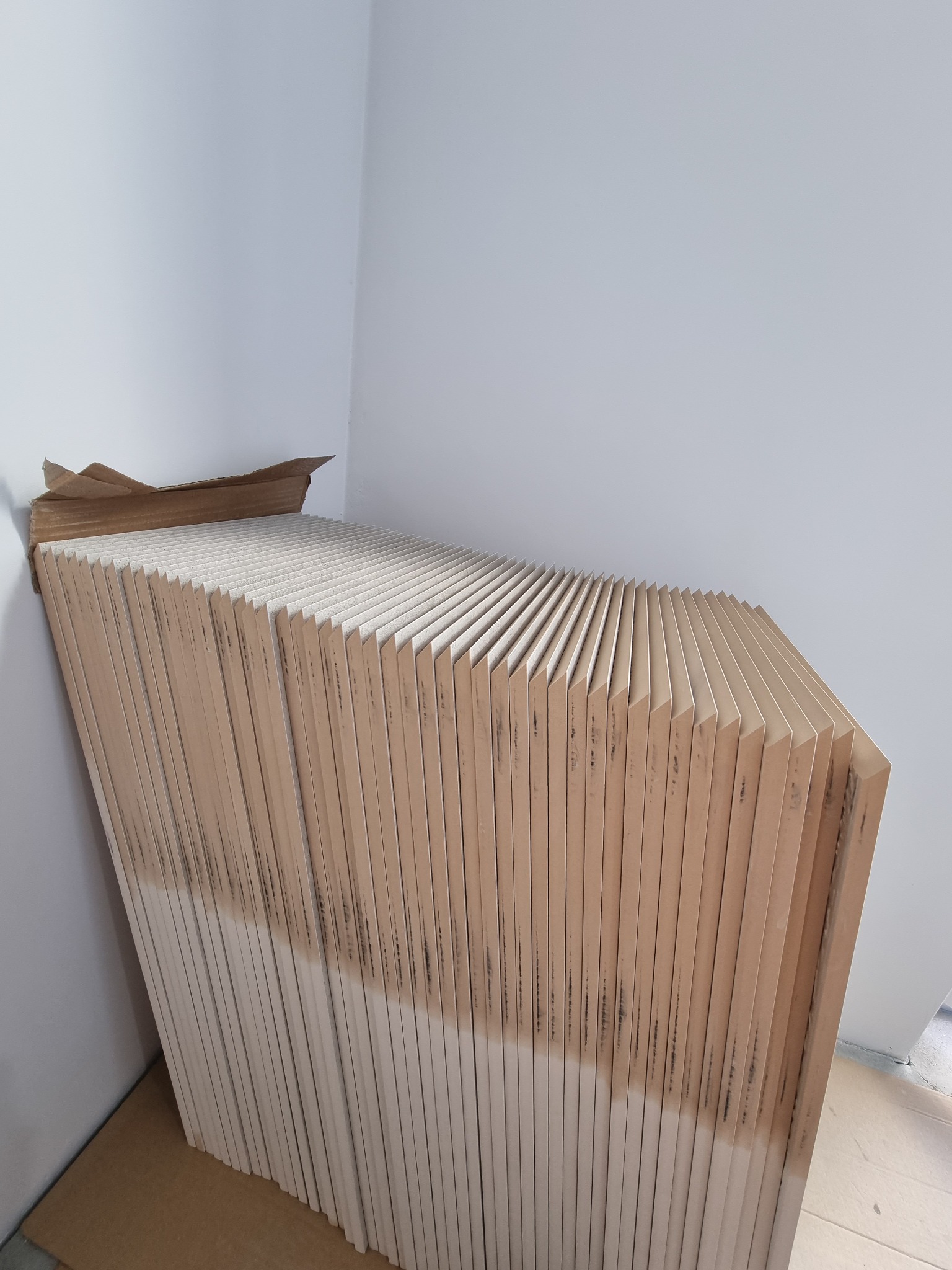 Stos płyt kartonowo-gipsowych w kartonowym opakowaniu, ustawionych pod ścianą, widoczne ślady zabrudzeń na dolnej części płyt.