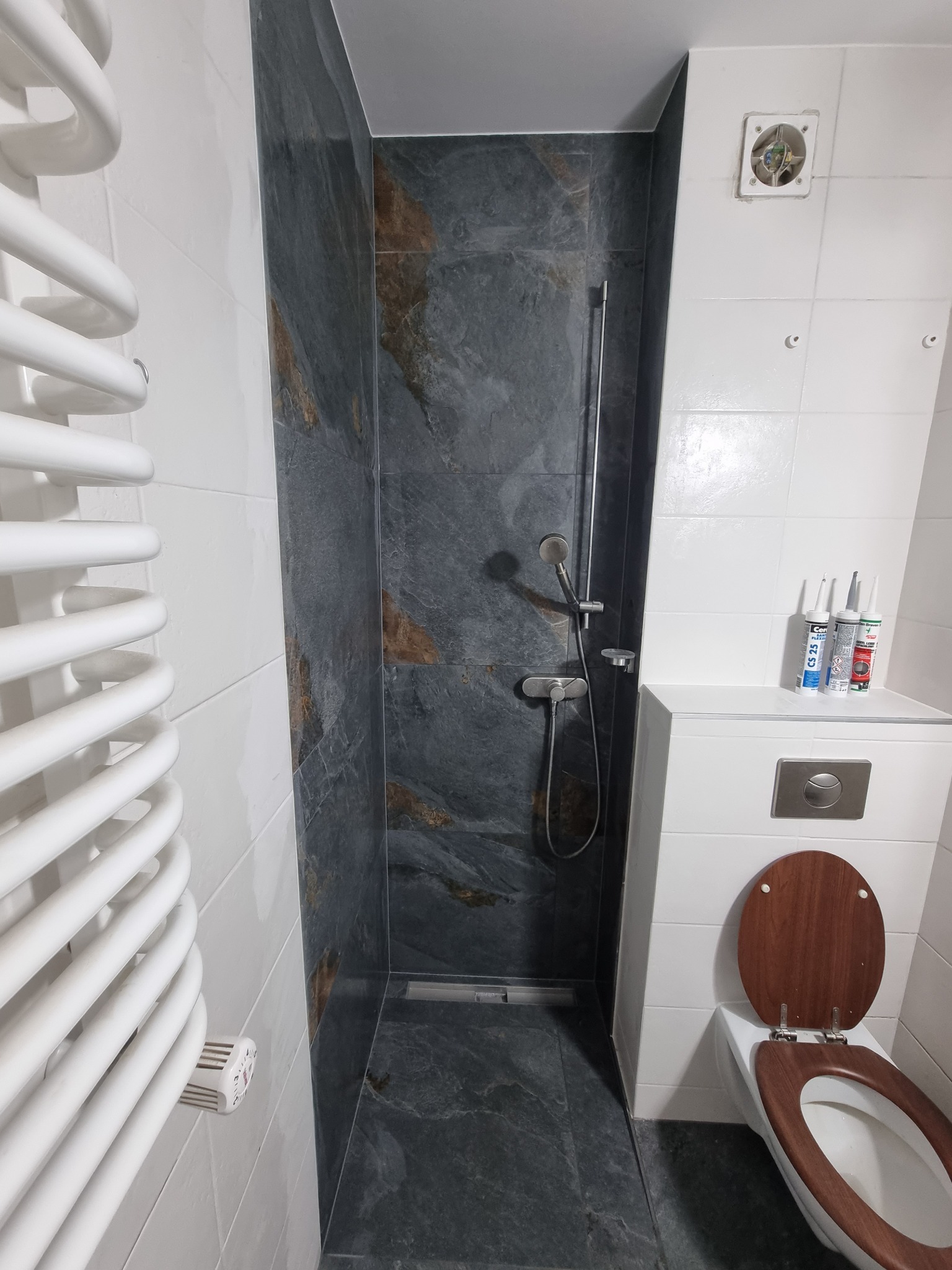 Nowoczesna kabina prysznicowa z szarymi płytkami imitującymi kamień, białe płytki na ścianach, grzejnik drabinkowy i toaleta z drewnianą deską. Na półce tuby z silikonem.