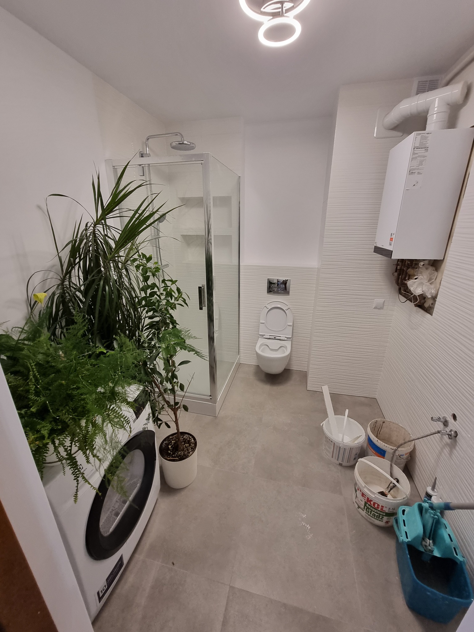 Wnętrze łazienki w trakcie prac wykończeniowych, widoczna kabina prysznicowa, toaleta, pralka z roślinami doniczkowymi obok oraz narzędzia i pojemniki budowlane na podłodze, ściana z dekoracyjną...