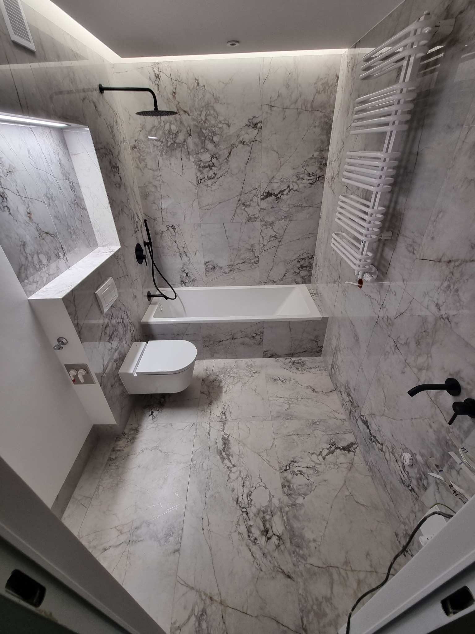 Nowoczesna łazienka wykończona marmurowymi płytkami, z podwieszaną toaletą, białą wanną, czarną armaturą i grzejnikiem drabinkowym.