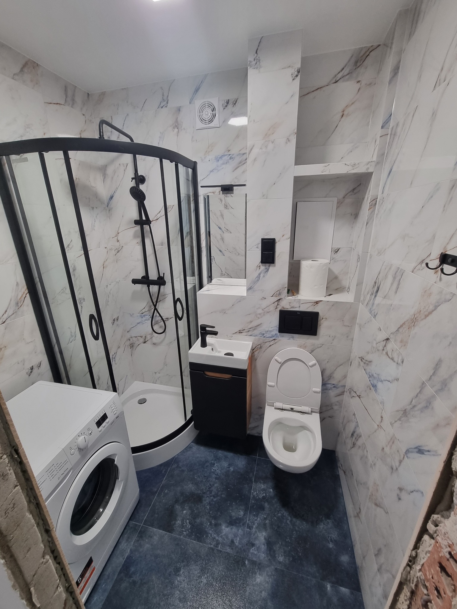 Wykończona łazienka z kabiną prysznicową z czarnymi profilami, umywalką nablatową na szafce, toaletą i pralką; marmurowe płytki na ścianach, ciemna podłoga.