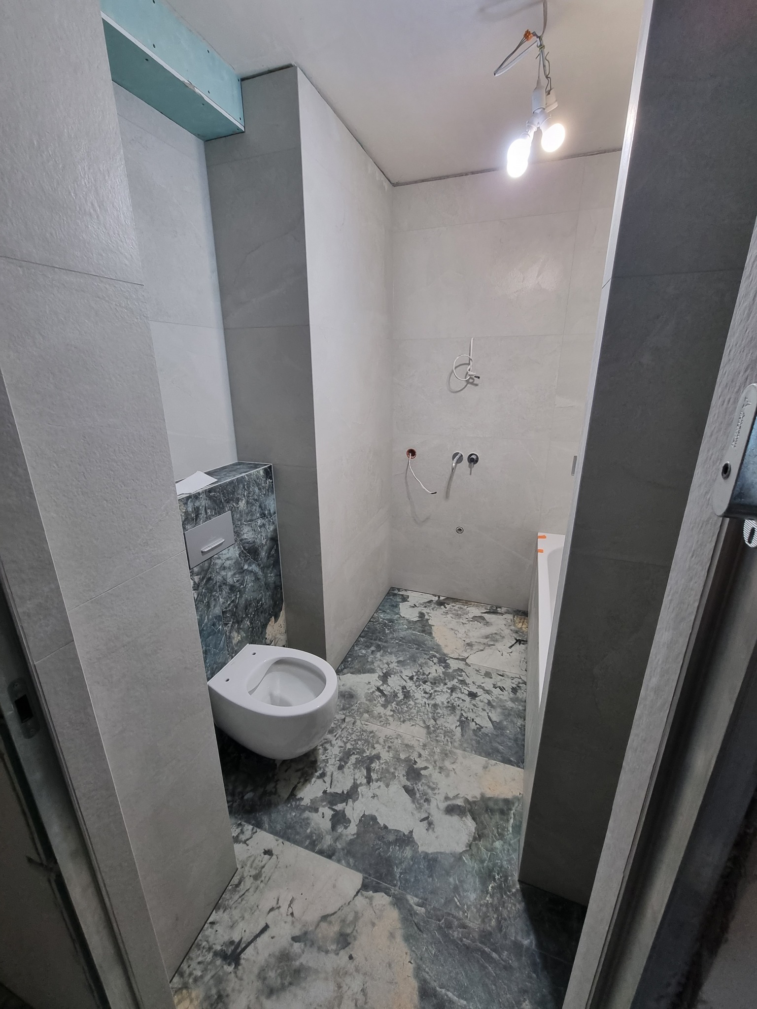 Wnętrze łazienki w trakcie remontu: szare płytki na ścianach, marmurowa podłoga, podwieszana toaleta z zabudową, widoczne instalacje wodne, surowa żarówka.
